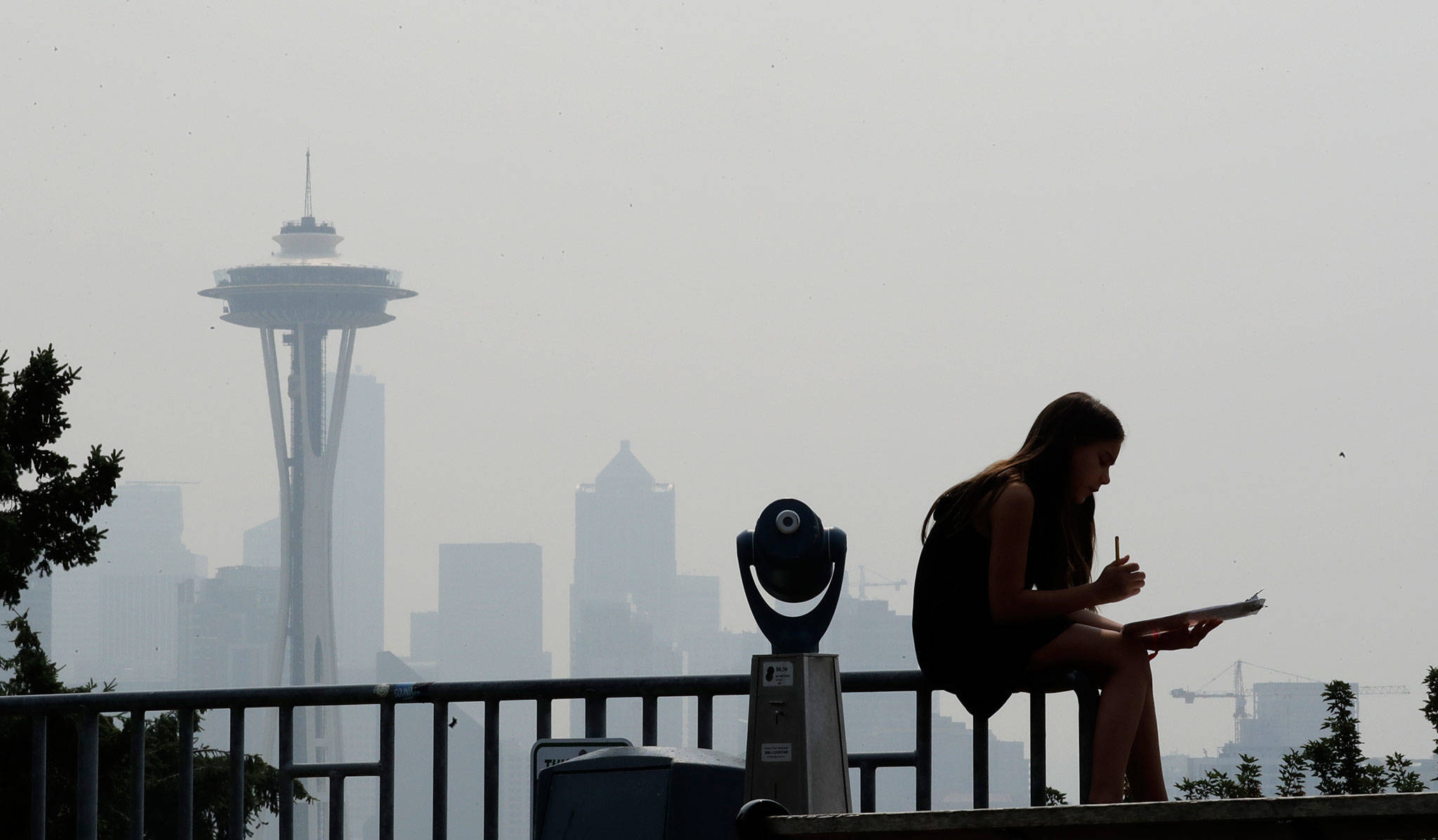 A woman works on a drawing next to an unused viewing scope as a smoky haze obscures the Space Needle and downtown Seattle last August as smoke from wildfires moved across the region. (Photo courtesy of The Herald/Elaine Thompson/Associated Press)